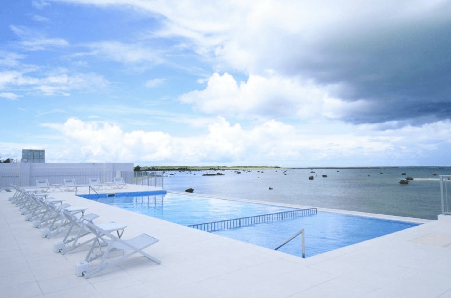 沖繩宮古島水印飯店 (Watermark Hotel Okinawa Miyakojima) 的戶外無邊際海景游泳池。純白色的池畔平台上整齊排列著一整排白色躺椅，泳池前方緊臨著佈滿天然礁石的寬闊淺灘與海洋，天空伴隨著藍天與雲朵。