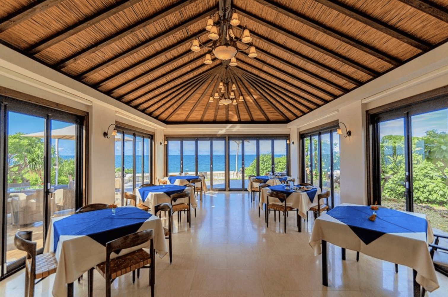 維拉布度假酒店 (Villabu Resort) 採光極佳的景觀餐廳，空間採用傳統茅草編織屋頂搭配古典吊燈，四周環繞著落地玻璃門窗，內部擺放數張鋪著亮藍色桌巾的餐桌，用餐時可飽覽戶外海景。