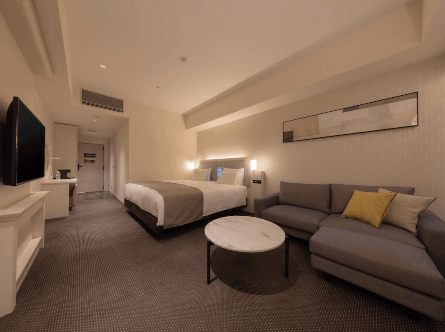 大阪LIBER酒店 (LIBER HOTEL OSAKA) 的寬敞客房,設有雙人床與大型的灰色L型轉角沙發,搭配圓形大理石紋茶几,牆面上掛有簡約的藝術畫作,空間充滿居家舒適感