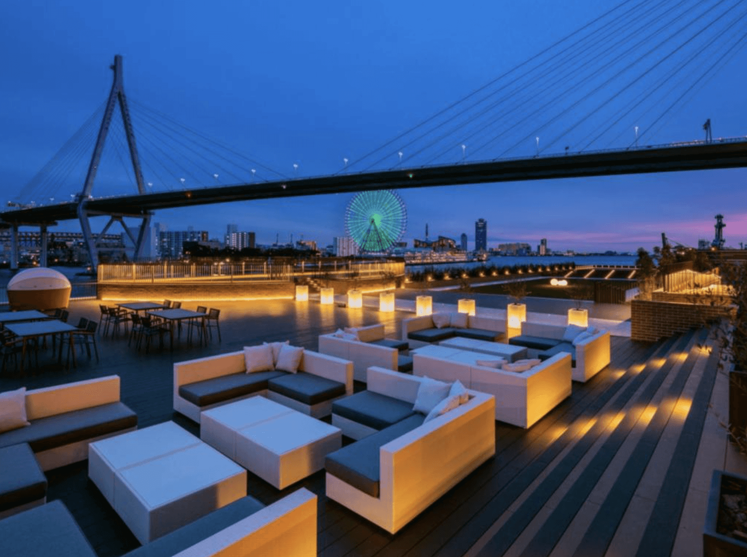 大阪LIBER酒店 (LIBER HOTEL OSAKA) 的戶外景觀露台,在傍晚的暖黃色燈光點綴下,設有多組白色的戶外休閒沙發座位,背景可飽覽壯觀的跨海大橋與點亮霓虹燈的摩天輪迷人夜景