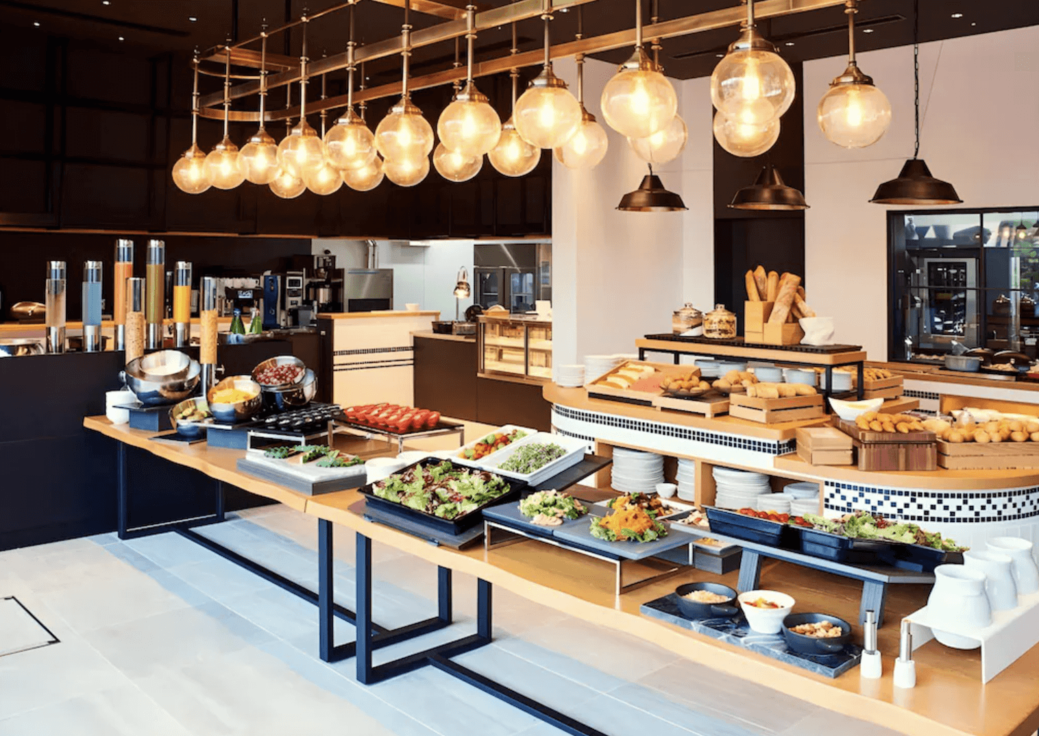 大阪LIBER酒店 (LIBER HOTEL OSAKA) 的明亮自助餐廳,長型的木質取餐檯上豐盛地陳列著各式新鮮沙拉、麵包與冷盤餐點,吧檯上方懸掛著一長排溫暖的球型玻璃吊燈,用餐環境溫馨且充滿質感