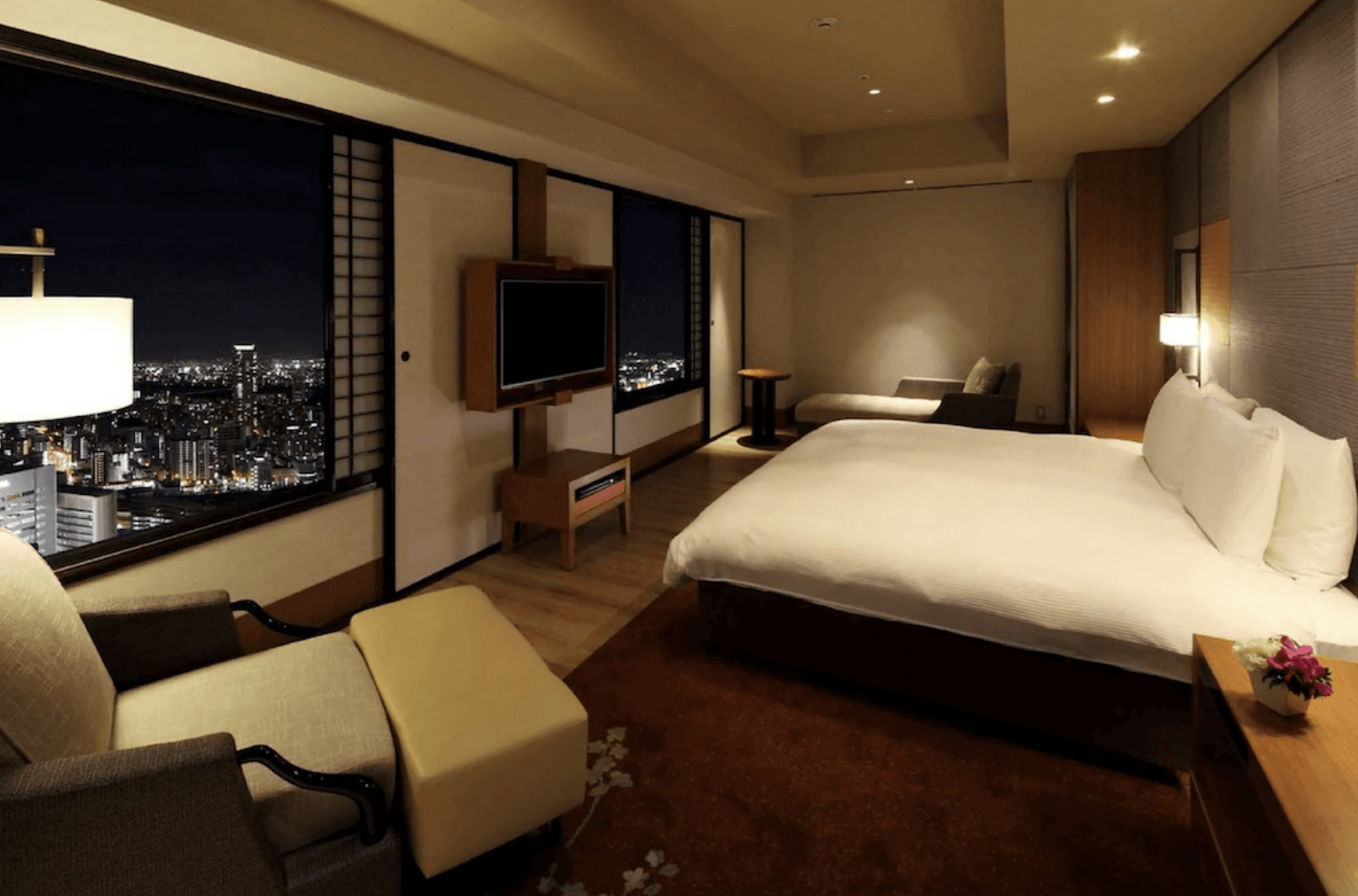 大阪希爾頓飯店客房的夜景。房間內有舒適的雙人床、壁掛電視、落地窗和一張扶手椅，窗外可以看到大阪明亮的城市夜景