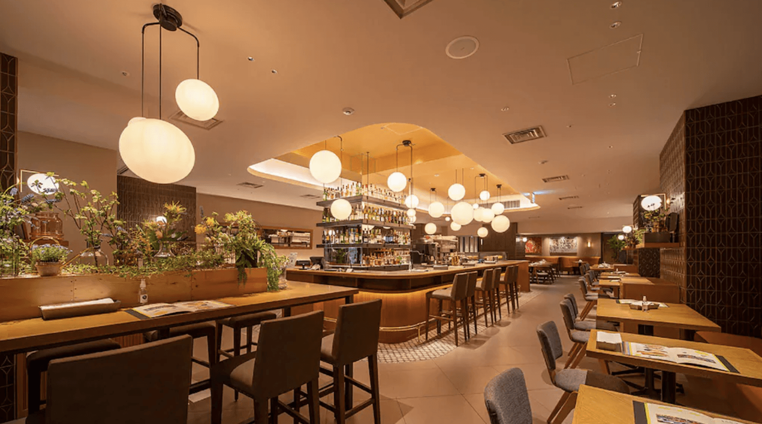 大阪格蘭比亞大飯店-JR大飯店集團的餐廳與酒吧空間，天花板懸掛著許多圓球造型吊燈，設有環形吧台座位區、木質餐桌椅以及點綴空間的綠色植物，氛圍溫馨且時尚