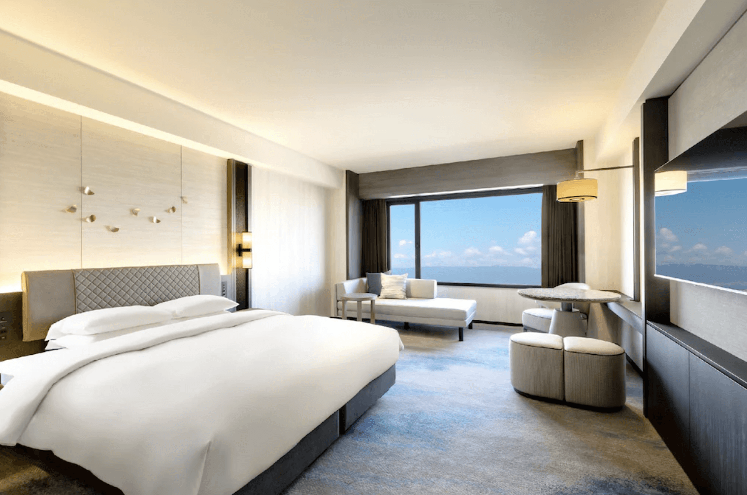 大阪灣格蘭王子飯店 (Grand Prince Hotel Osaka Bay) 的明亮海景雙人客房。房內配備一張舒適的大型雙人床、壁掛式電視,以及靠窗的淺色休閒長沙發與圓形茶几。大面景觀窗外可見晴朗無雲的藍天與廣闊的蔚藍海景,整體空間採光極佳,設計簡約現代
