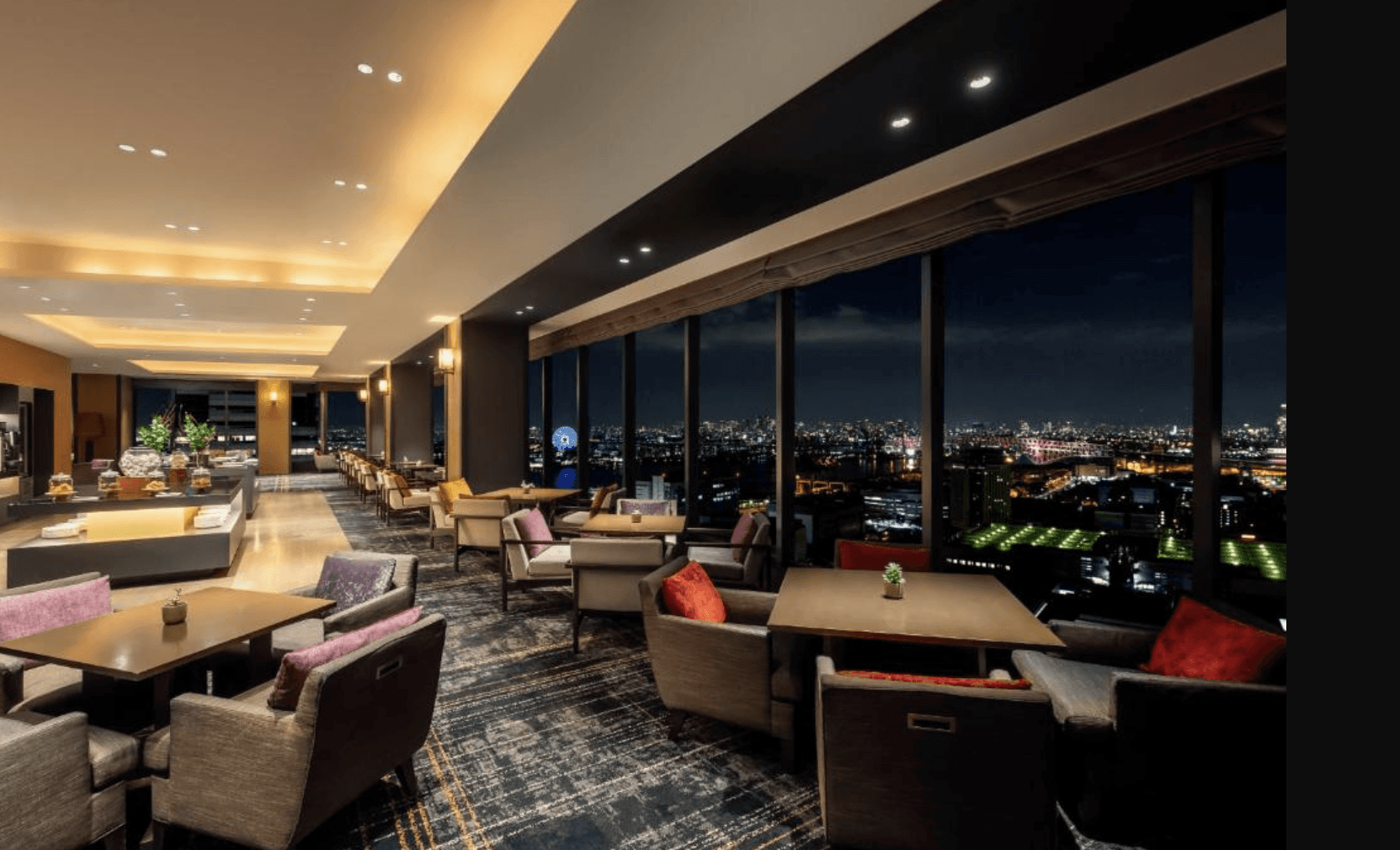 大阪灣格蘭王子飯店 (Grand Prince Hotel Osaka Bay) 的高樓層景觀餐廳與休息區。空間內擁有大面積的落地窗,能將璀璨絢麗的大阪城市夜景、發光的摩天輪與跨海大橋盡收眼底。室內鋪設深色地毯,設有多組舒適的沙發座椅、餐桌及後方的吧檯區,環境典雅奢華