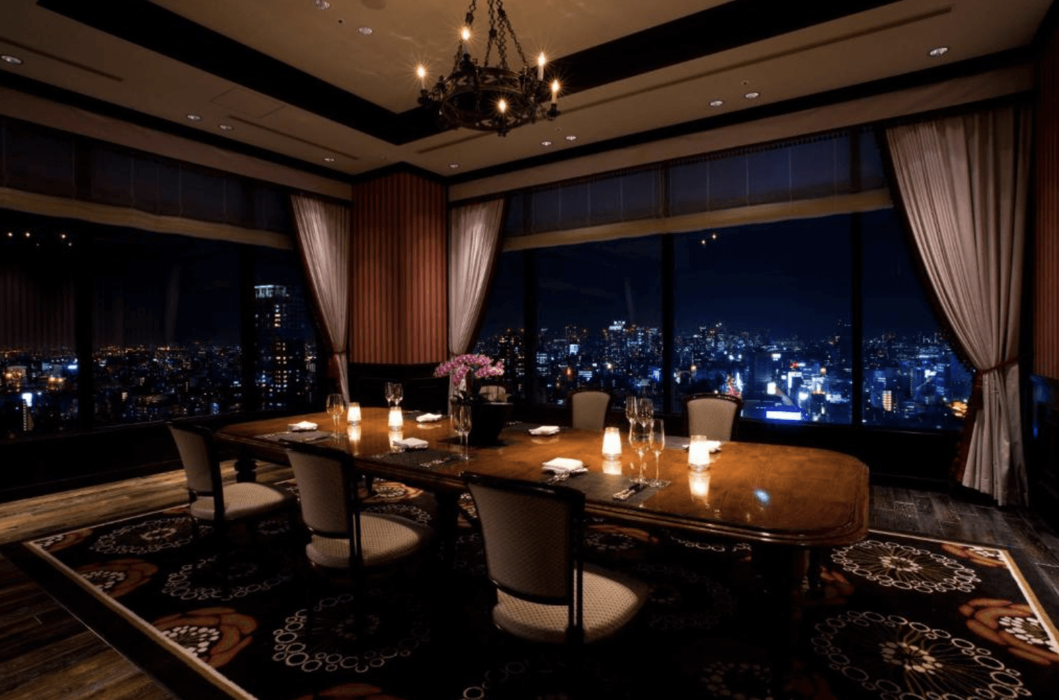 大阪蒙特利格拉斯米爾酒店的高空景觀餐廳包廂，空間內鋪設深色圖騰地毯，中央擺放著高雅的長型木質餐桌與餐椅，透過大片玻璃窗可將絢麗的城市夜景盡收眼底