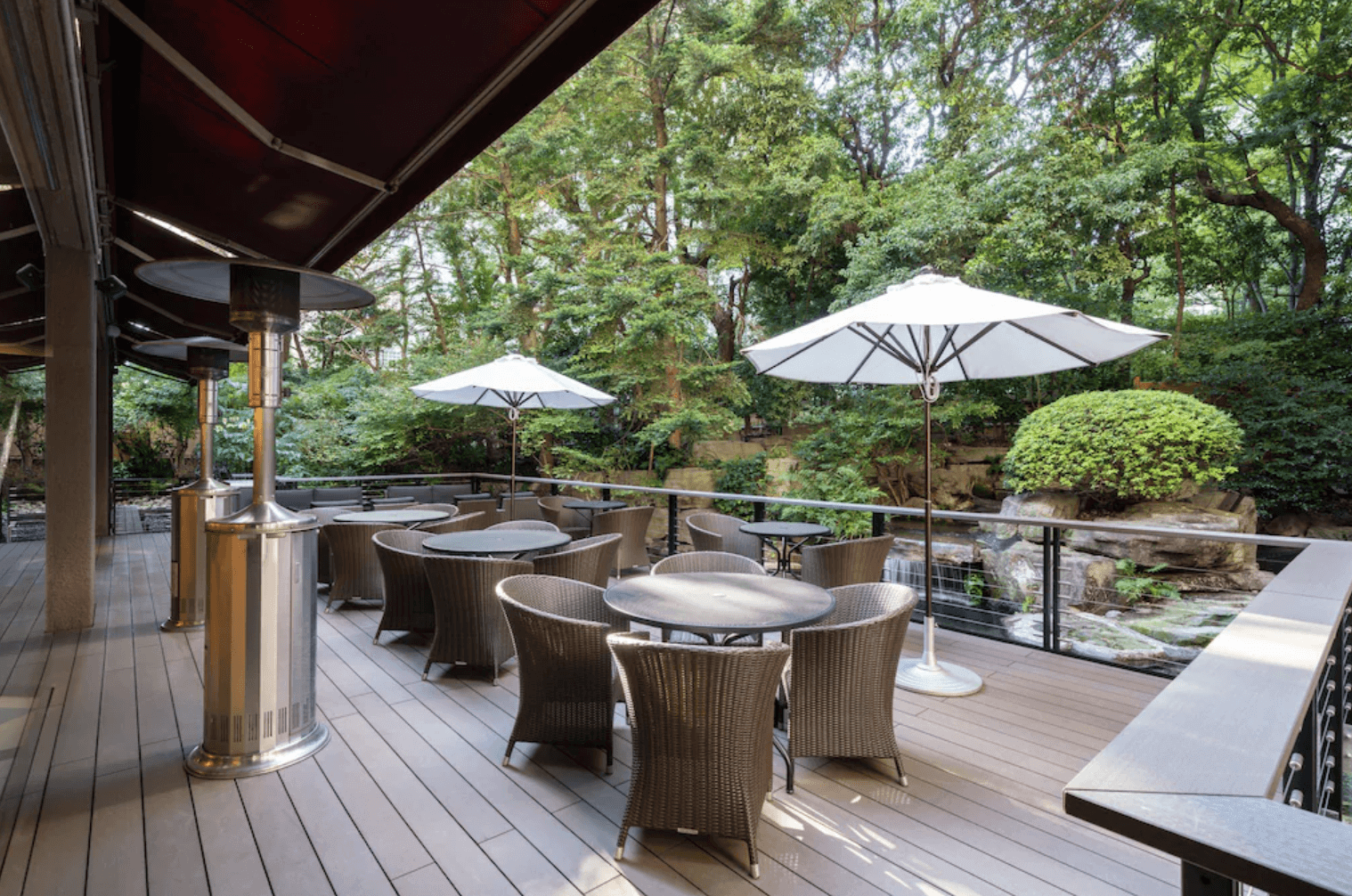 大阪麗嘉皇家飯店-洲際酒店集團Vignette系列的戶外露台休息區，木質地板上擺放著多組戶外編織座椅與圓桌、白色遮陽傘及直立式暖爐，周圍環繞著翠綠的樹木與庭園水池造景