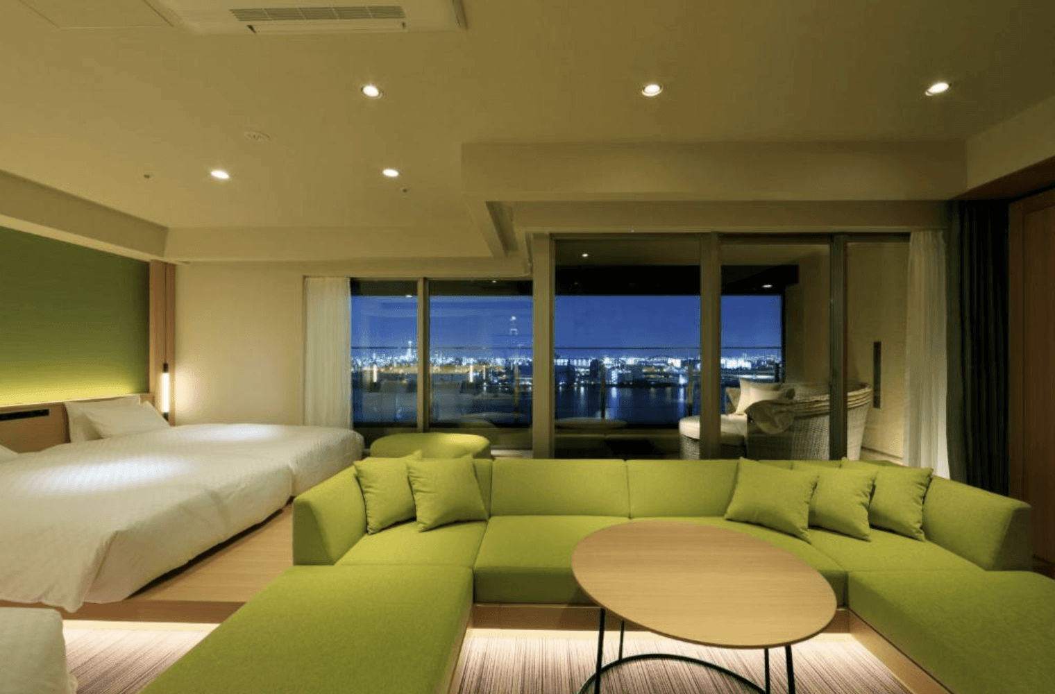 日本環球影城新格拉利飯店及空中水療中心 (The Singulari Hotel & Sky Spa at Universal Studios Japan) 的寬敞景觀客房,木質架高地板上設有兩張大型床位,前方搭配舒適的綠色L型大沙發與圓形茶几,落地窗外展現迷人的水岸城市夜色