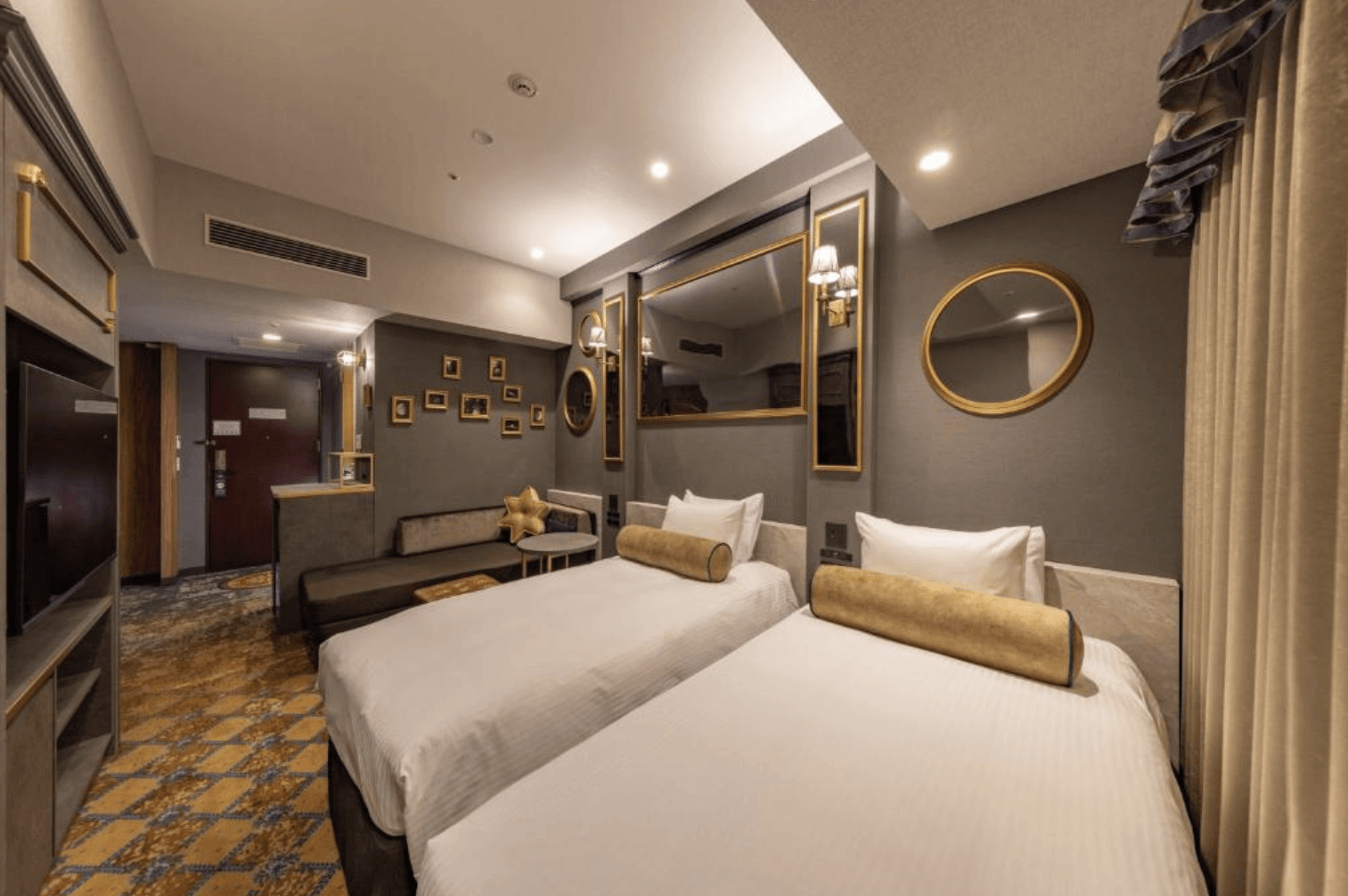 環球大廈京阪飯店 (Hotel Keihan Universal Tower) 的優雅雙人客房,牆面以沉穩的深灰色搭配金色邊框的鏡面與相框裝飾,兩張單人床配有精緻的金色圓柱形抱枕,角落設有舒適的深色沙發休息區