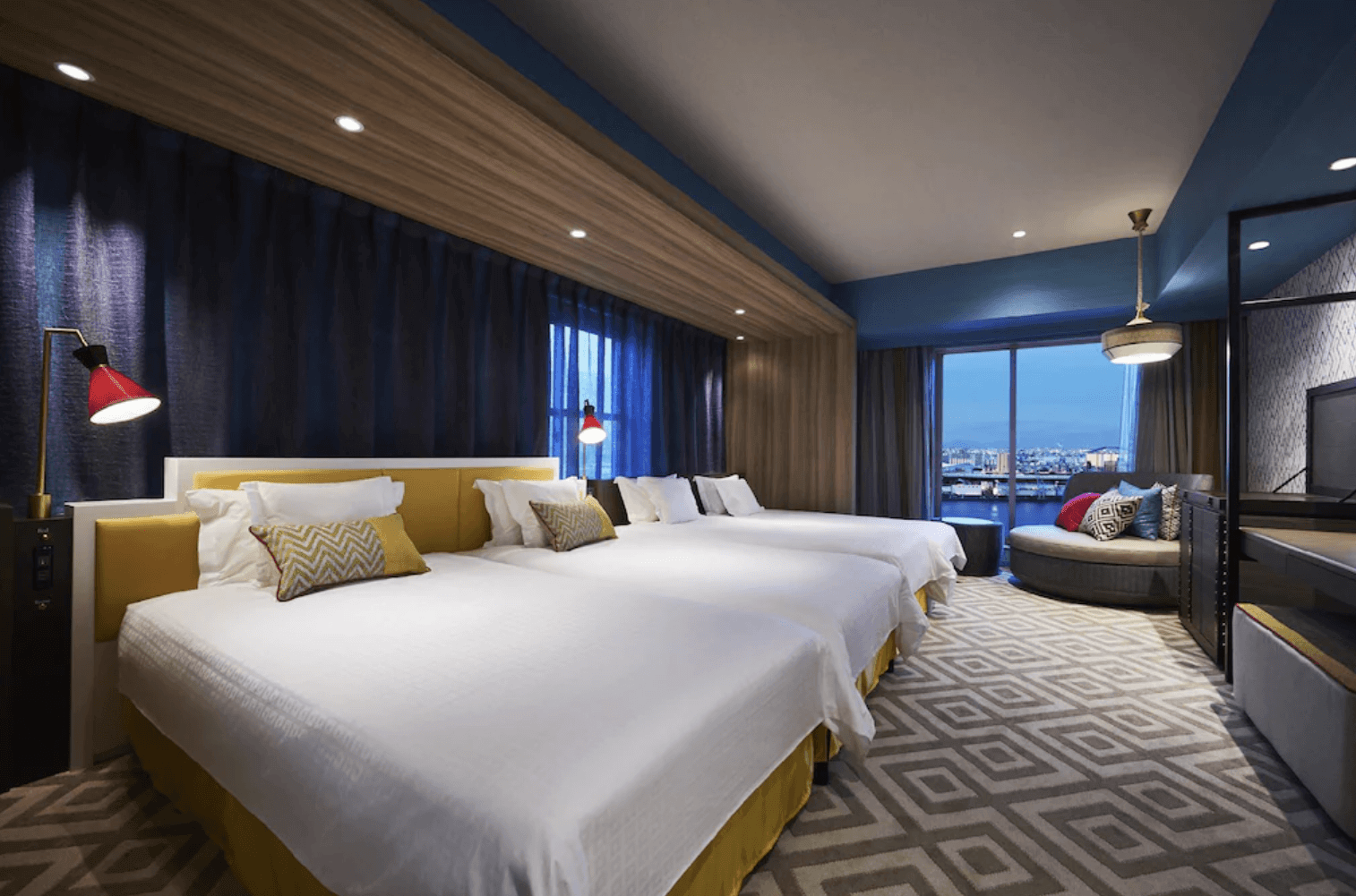 環球影城港灣酒店 (Hotel Universal Port) 的海景三人客房,房內並列著三張單人床,配有深藍色窗簾與角落的圓形休閒沙發,大面窗戶可眺望傍晚的港灣景色