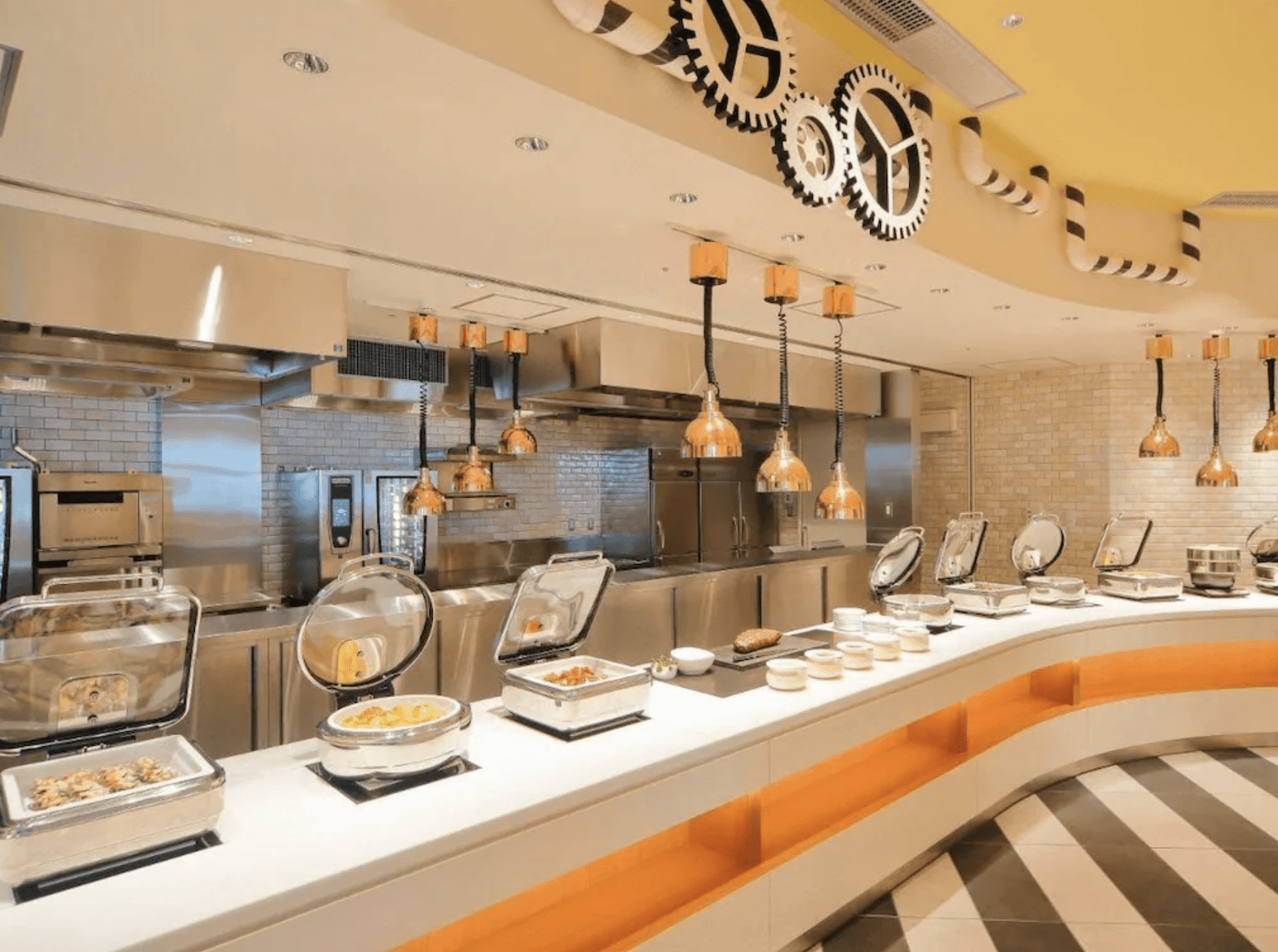 環球影城維塔港酒店 (Hotel Universal Port Vita)的自助餐廳取餐區,明亮的白色弧形吧檯上陳列著多組保溫餐爐,上方懸掛著金屬質感保溫燈,天花板帶有獨特的齒輪造型裝飾