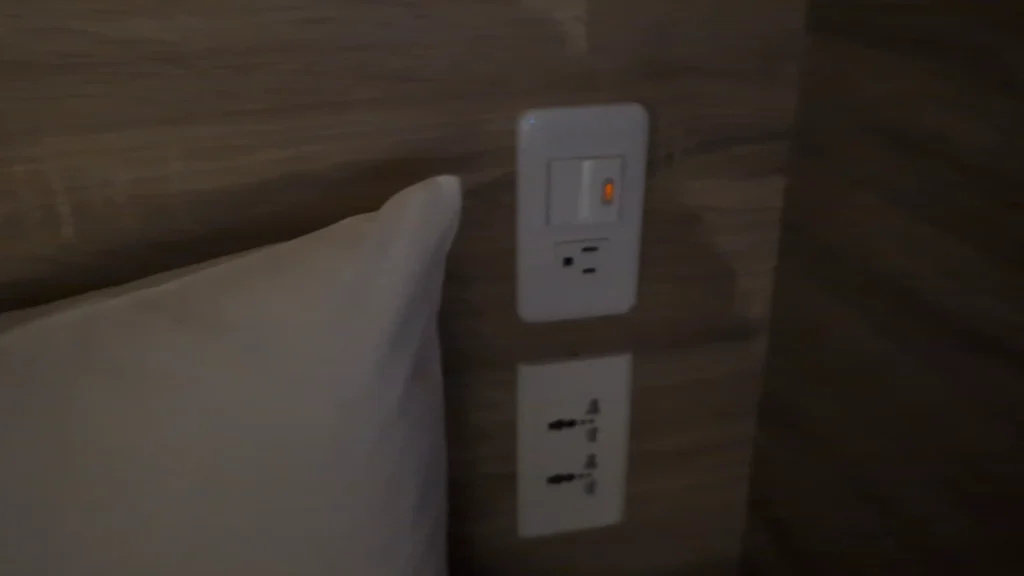 木質床頭背板旁的白色電源面板特寫，上方為附指示燈的電燈開關與標準三孔插座，下方則配有兩個可兼容多國規格的萬用電源插座