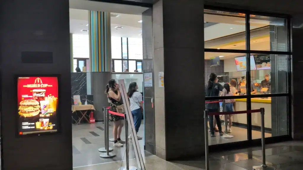 黑色金屬玻璃門窗隔出的麥當勞餐廳，入口處立有排隊紅龍柱與「FIFA WORLD CUP」漢堡套餐亮面燈箱海報，室內點餐櫃檯前有多位顧客正在等候點餐