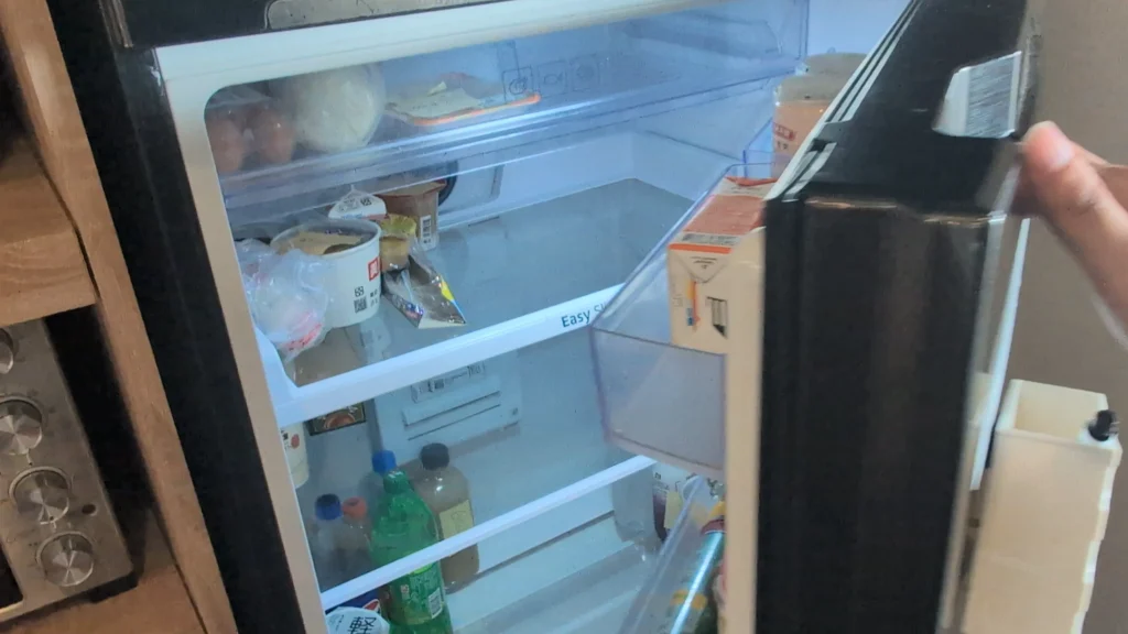 一隻手打開的共用雙門冰箱冷藏室，透明層架上存放著碗裝優酪乳、紙盒包裝食品與瓶裝飲料，側門附有筆記欄與分類置物槽