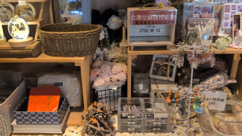 一個鋪著鳳梨圖案桌布的木質分層陳列櫃，擺滿了各種雜貨和手工藝品。上層有編織籃、乾燥花、發光瓶、誠實箱，下層有貼著「開工大吉」的紅包、化妝包、亞克力收納盒，以及掛滿鑰匙圈（標價60元）的展示架