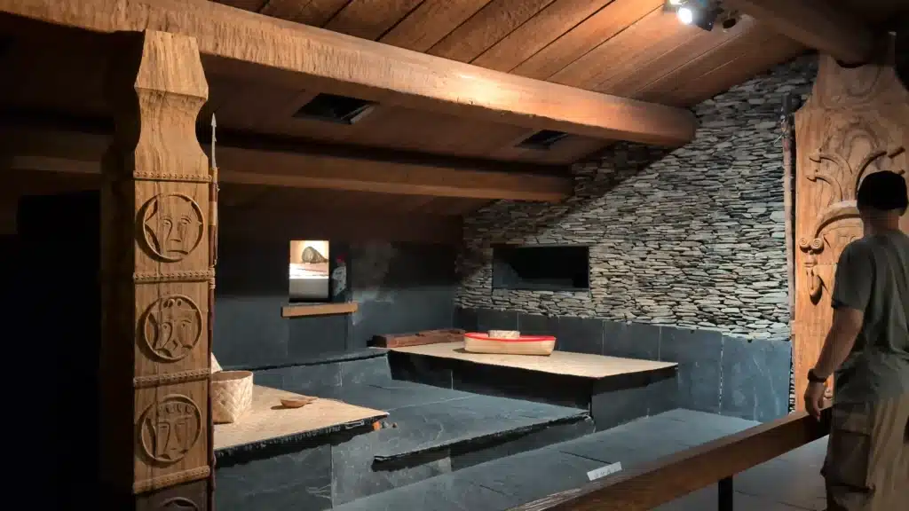 昏黃燈光下的排灣族或魯凱族傳統石板屋室內展區，包含厚重木樑屋頂、疊石板岩牆面、抬高的石板床鋪與編織器皿，左側立有雕刻人面紋樣的原木圖騰柱