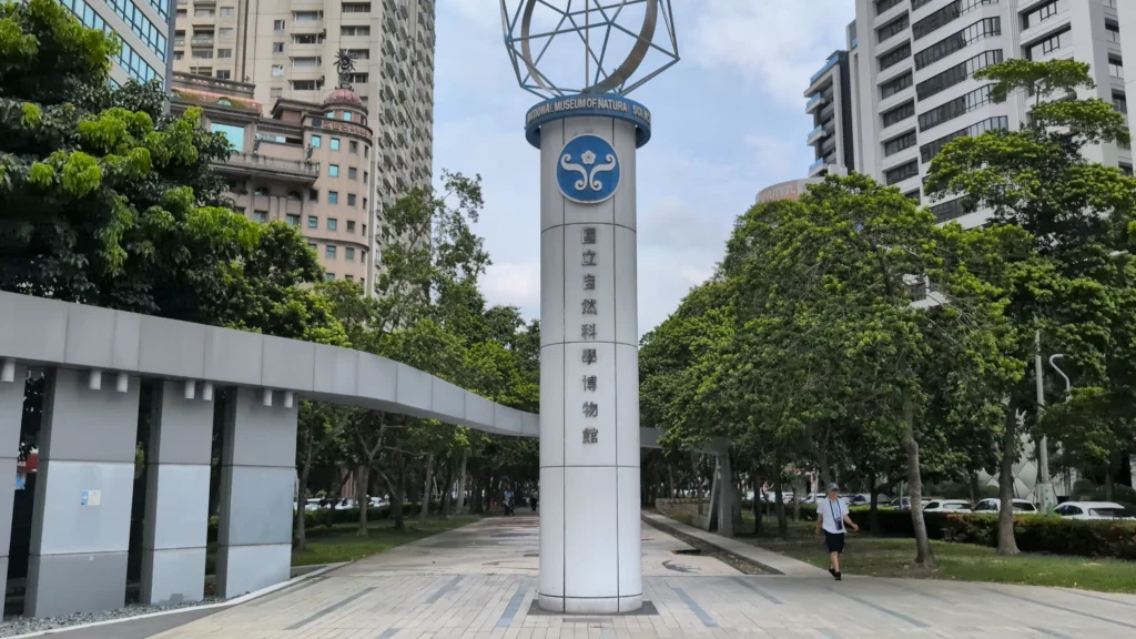 台中科博館外的標誌性白色圓柱地標，上方刻有館徽與「國立自然科學博物館」字樣，頂部設有幾何球形裝飾，背景為林蔭步道、現代建築與步行的行人