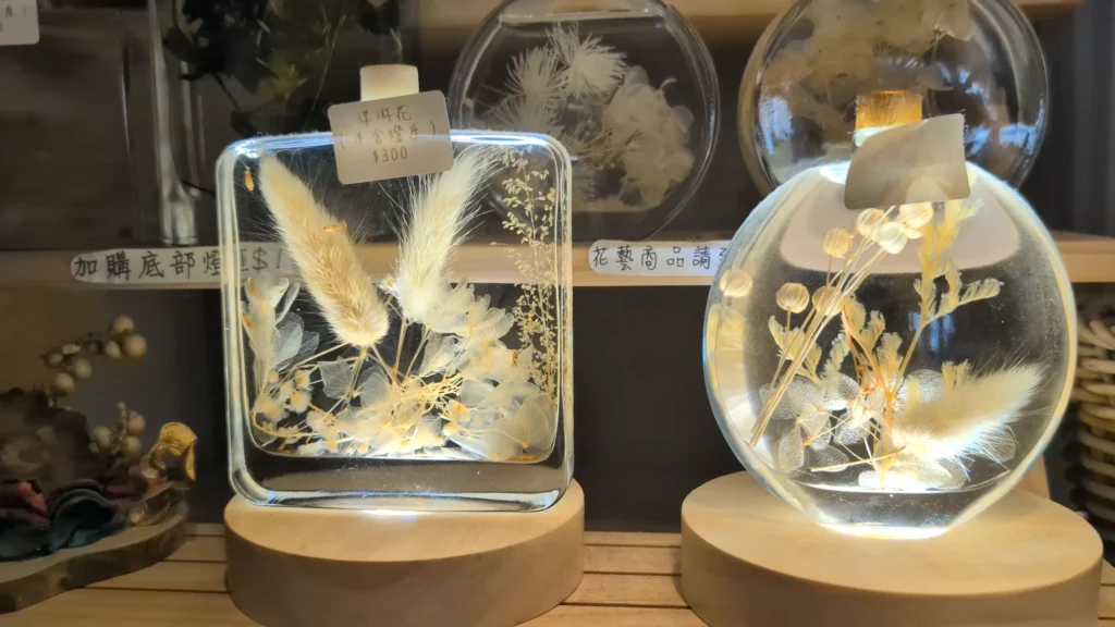 在木製發光燈座上展示的兩瓶浮游花。左側是方形瓶，內含白色和米色的乾燥羽毛狀與團狀植物，貼有「浮游花（不含燈座）$300」標籤。右側是圓形瓶，內含米色乾燥花和果實