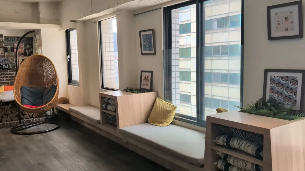 採光充足的室內大廳空間，地面鋪設深灰色木紋地板。靠窗處設計了一整排木質臥榻，上方擺放淺色座墊與黃色靠枕，並結合了兩座附有收納軟墊與綠色植栽的矮櫃。左側角落放置一張蛋形藤編吊椅，牆面掛有多幅卡通插畫與復古裝飾畫，窗外可見對面的現代建築外牆