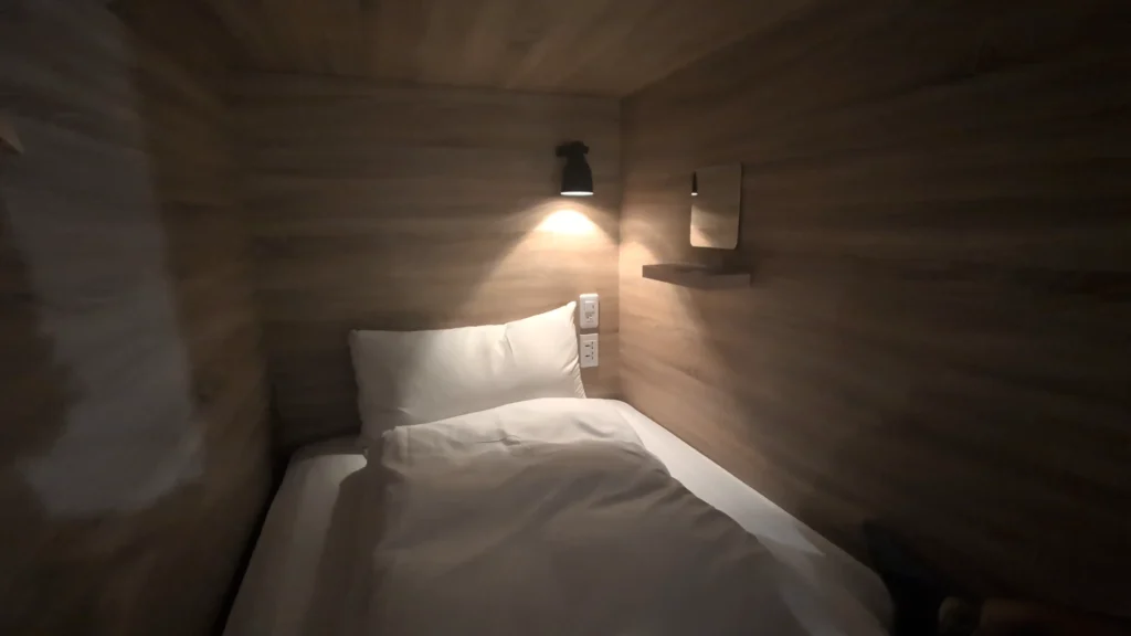 暖黃光照映下的單人膠囊床位全景，鋪有白色床單與蓬鬆枕頭被褥，木質牆面上設有一盞黑色壁燈、小型梳妝鏡、置物層板及多組電源插座