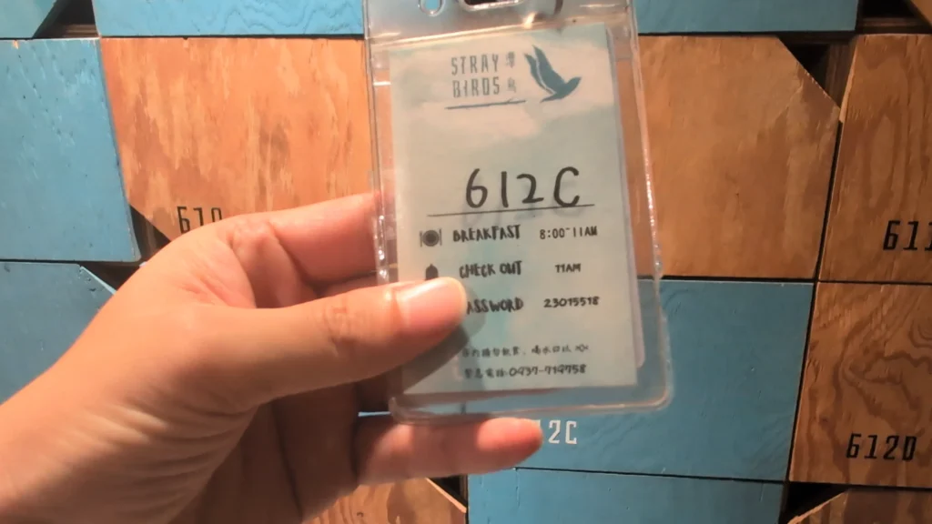 手持「STRAY BIRDS 漂鳥」612C 號透明房卡，卡片上印有早餐時間與 Wi-Fi 密碼等資訊，背景為標示著相對應房號的藍色與木紋拼色收納櫃