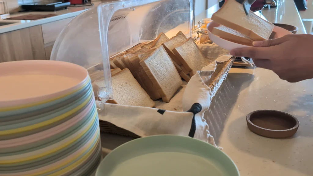 自助吧台上的透明掀蓋防塵麵包盒，裡面整齊排滿切片白吐司，一旁堆疊著彩色餐盤，房客正用食物夾夾取吐司放入盤中