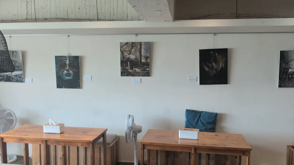 挑高且簡約的室內用餐空間，白色牆面懸掛五幅不同風格的掛畫，下方擺放兩張長方形木質餐桌、木椅、靠墊與站立式電風扇