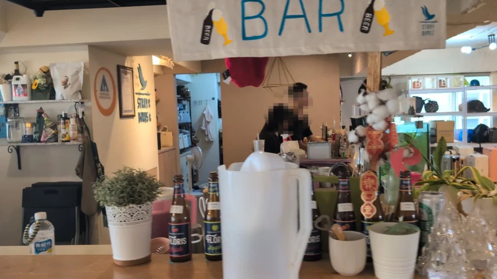室內吧檯與櫃台區域，上方掛著手繪風格的「BAR」掛布與啤酒圖案，木質吧檯桌面上擺放著多瓶水果啤酒、綠色盆栽與冷水壺，後方有工作人員與充滿綠意的陳列架