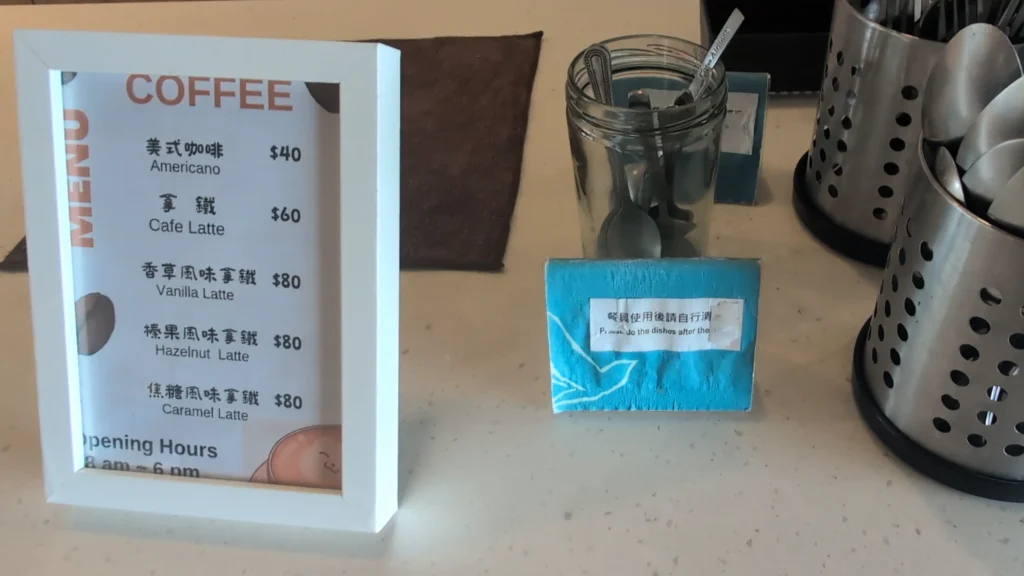 廚房吧台上的白框咖啡菜單看板。看板上寫著：美式咖啡 $40、拿鐵 $60、香草風味拿鐵 $80、榛果風味拿鐵 $80、焦糖風味拿鐵 $80。旁邊有放餐具的玻璃罐和金屬筒