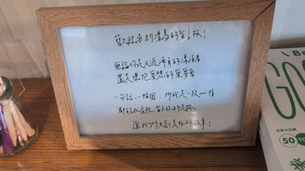 木框相框中展示的手寫文字卡，內容為「歡迎來到漂鳥的留言板！無論你是天涯海角的漂泊者，還是懷抱夢想的築夢者……讓我們共創美好的故事！」，旁邊放置著彩色筆筒