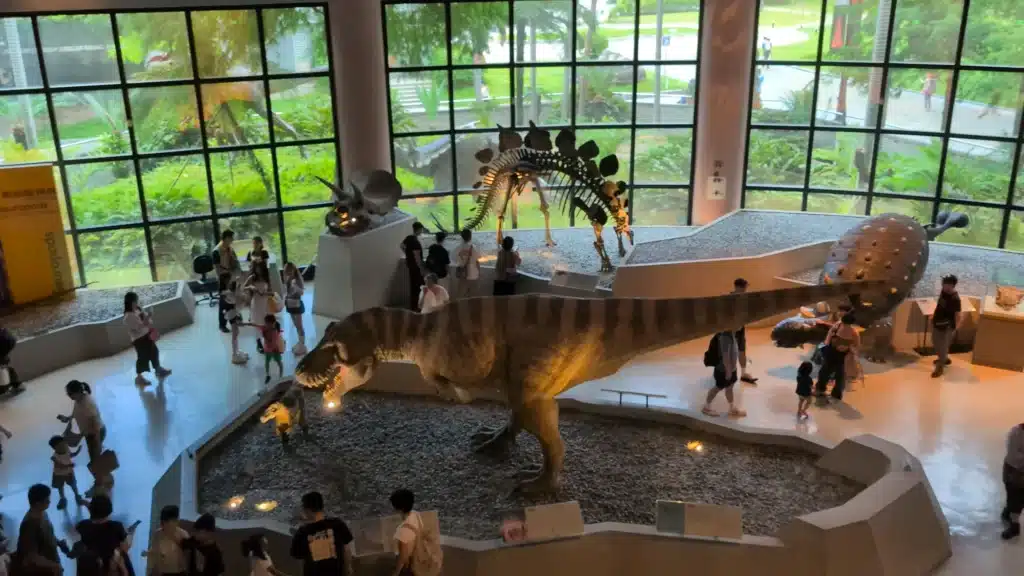 高挑明亮的恐龍展示廳，大片落地窗外映出戶外綠意。展區碎石台上擺設著大型機械暴龍、劍龍化石骨架及甲龍模型，步道上有許多家長帶著孩童圍繞觀賞
