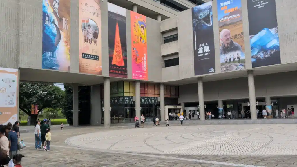 科博館主建築正面視角，灰色石材外牆上高掛著「海龍王滄龍」、「珍古德」等多幅大型展覽宣傳垂簾，下方開闊的圓形紋理地磚廣場上有許多遊客與親子漫步