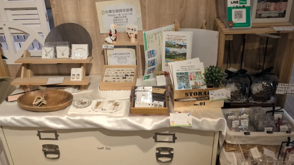 一個鋪著白色布、擺滿各式雜貨的白色工作櫃台。左側木架展示著項鍊與耳環，櫃台中央有放髮夾的圓形木盤、戒指盒、印有「此攤位錢錢投這裡」的告示、以及多本旅遊手冊。右側有 Line Pay 付款碼與禮盒包裝