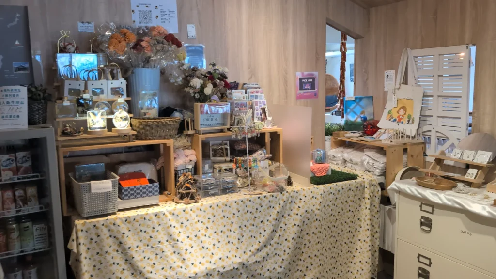 一間充滿木質元素的溫馨小店內部。左側是玻璃門冰箱，右側陳列著鋪有鳳梨圖案桌布的長桌，桌上擺滿了乾燥花、手工藝品、鑰匙圈和收納盒。靠牆有帆布袋、明信片架和飾品架