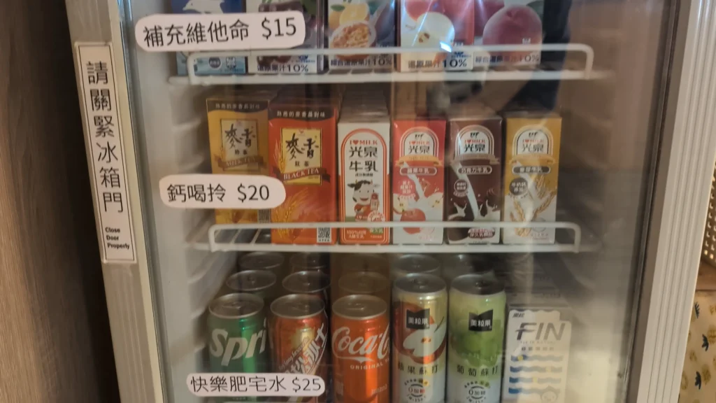 一個玻璃門冰箱，整齊陳列著各式飲料。上層是利樂包飲料（如麥香、光泉牛乳），貼有「補充維他命 $15」和「鈣喝拎 $20」手寫標籤。下層是罐裝飲料（如雪碧、可樂、FIN、美粒果），貼有「快樂肥宅水 $25」標籤