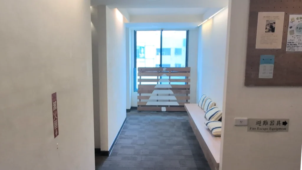 鋪設灰色地毯的客房走廊，白色牆面貼有避難器具指示，右側設有擺放靠枕的木製長椅與佈告欄，走廊底端落地窗前擺設了以木棧板彩繪的聖誕樹裝飾