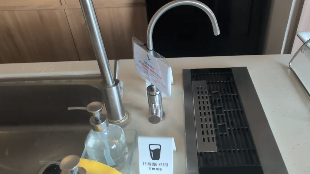 廚房水槽特寫。中央是一個弧形的可飲用水水龍頭，上面掛著一個牌子。水龍頭前放著一個白色標示牌，寫著：DRINKING WATER 可飲用水。左邊是廚房水龍頭、洗手乳和菜瓜布