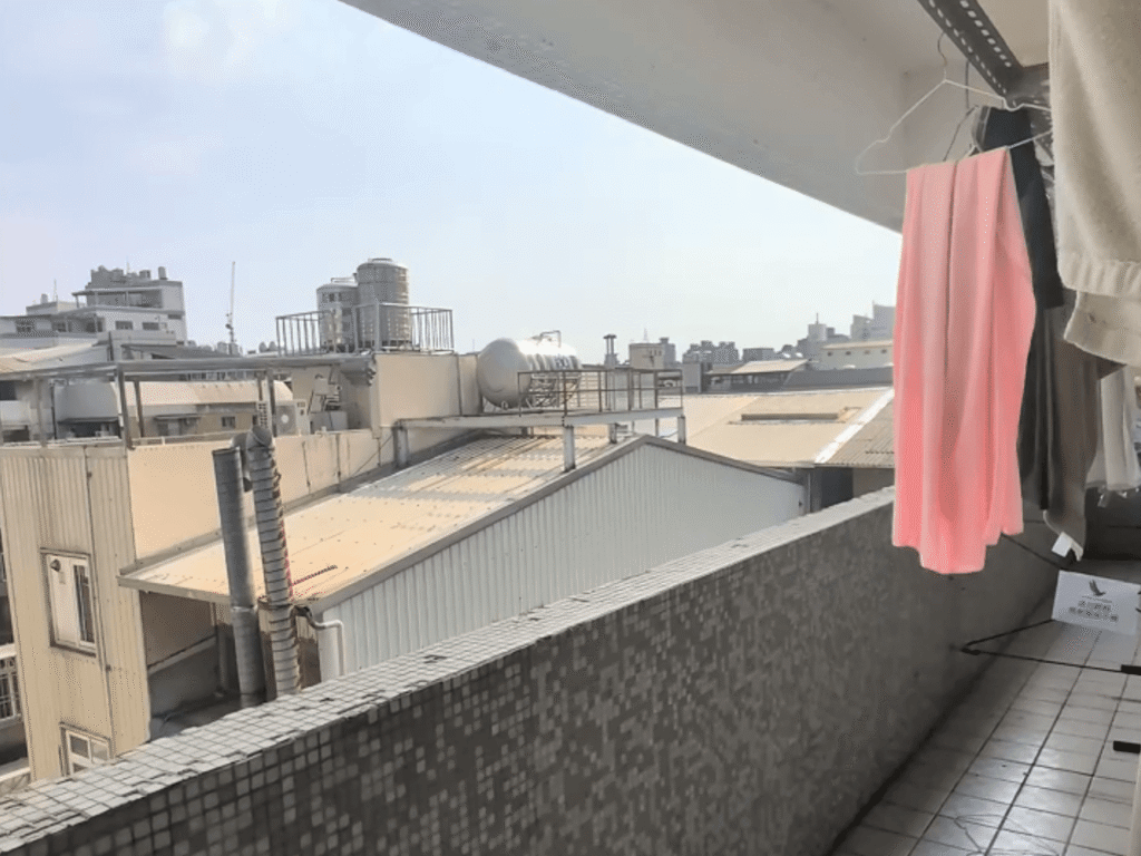 草悟道漂鳥青年旅館鋪設磁磚的半開放式長條陽台，欄杆為灰色小馬賽克磁磚，上方曬衣架掛著粉色衣物與毛巾，遠處可俯瞰鄰近建築的鐵皮屋頂、水塔及城市天空景觀